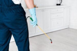 cropped view of exterminator in uniform holding spray with pesticide near kitchen cabinet